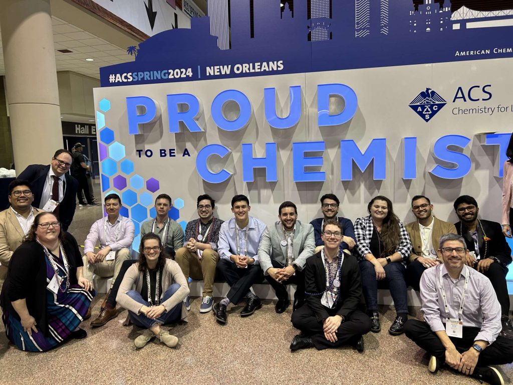 A group of professionally dressed folks which include Merck employees, PROF leaders, and the 2024 ACS Pride Merck Graduate Research Award recipients. They are all sitting or posing right in front of the ACS's "Proud to be a Chemist" signage.