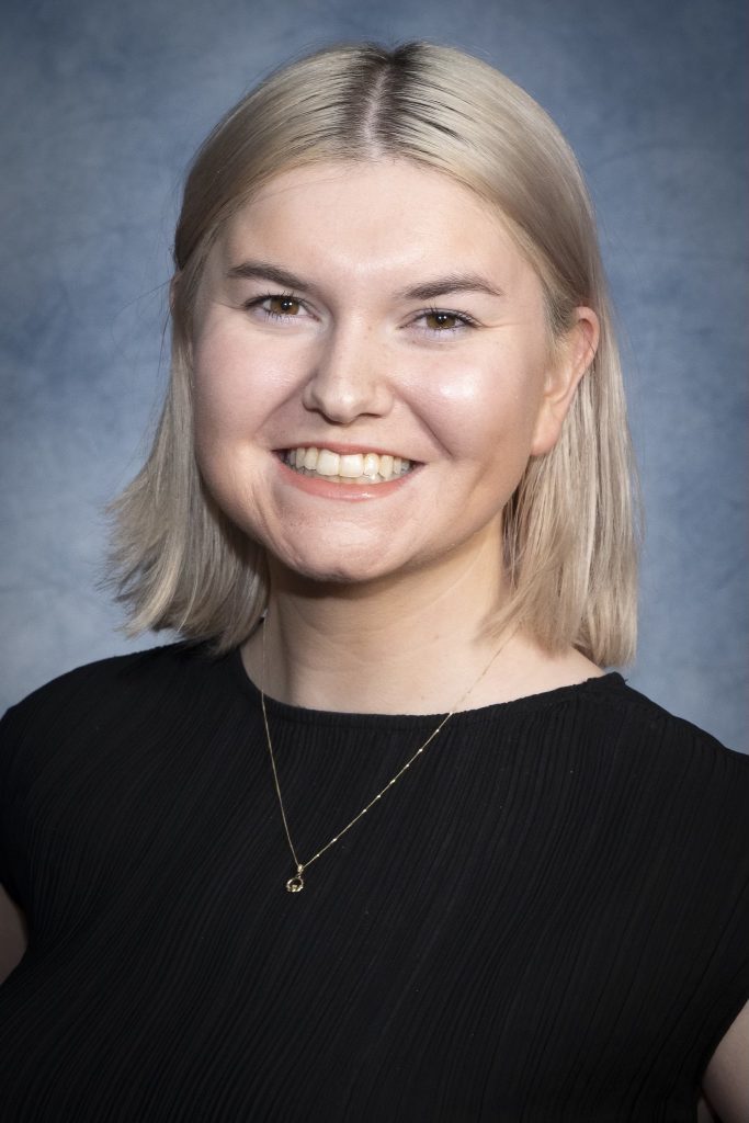 A white woman with blonde shoulder length hair in a black top and a thin chained necklace smiling back at the camera.