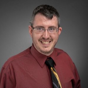 A dark-haired man with glasses in a red button up and black and yellow tie