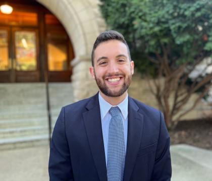 A man in a navy blue suit jacket and light blue dress shirt and tie grinning in front of the steps of a building.