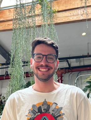 A man with brown hair and cat eye glasses in a T-shirt with the New York City skyline in front of vines.