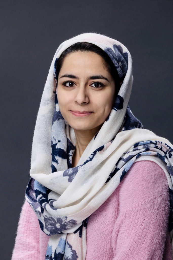 A woman with a white with blue floral head covering and a light pink long sleeved shirt facing the camera.