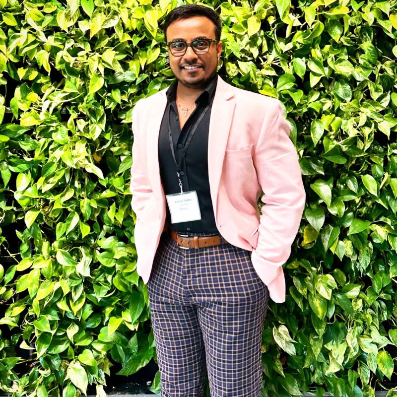 A dark-skinned man in a light pink business suit with dark dress shirt and patterned slacks in front of greenery.